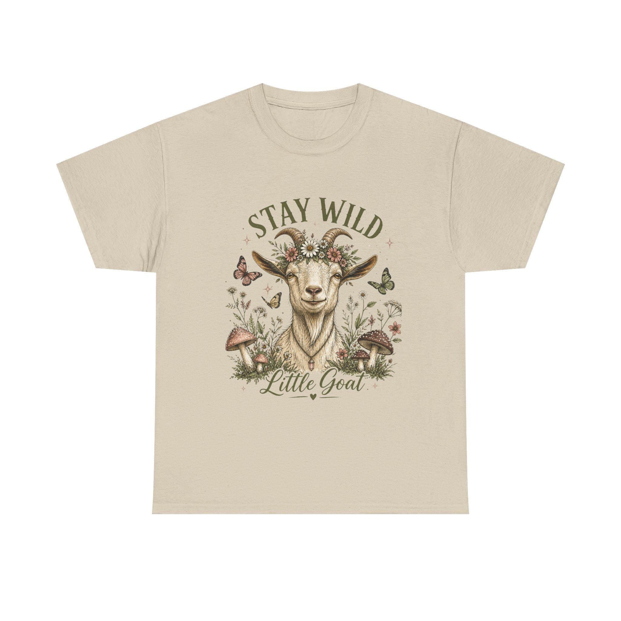 A light beige t-shirt with a graphic of a goat wearing a floral crown, surrounded by mushrooms and butterflies, with the text "Stay Wild Little Goat."