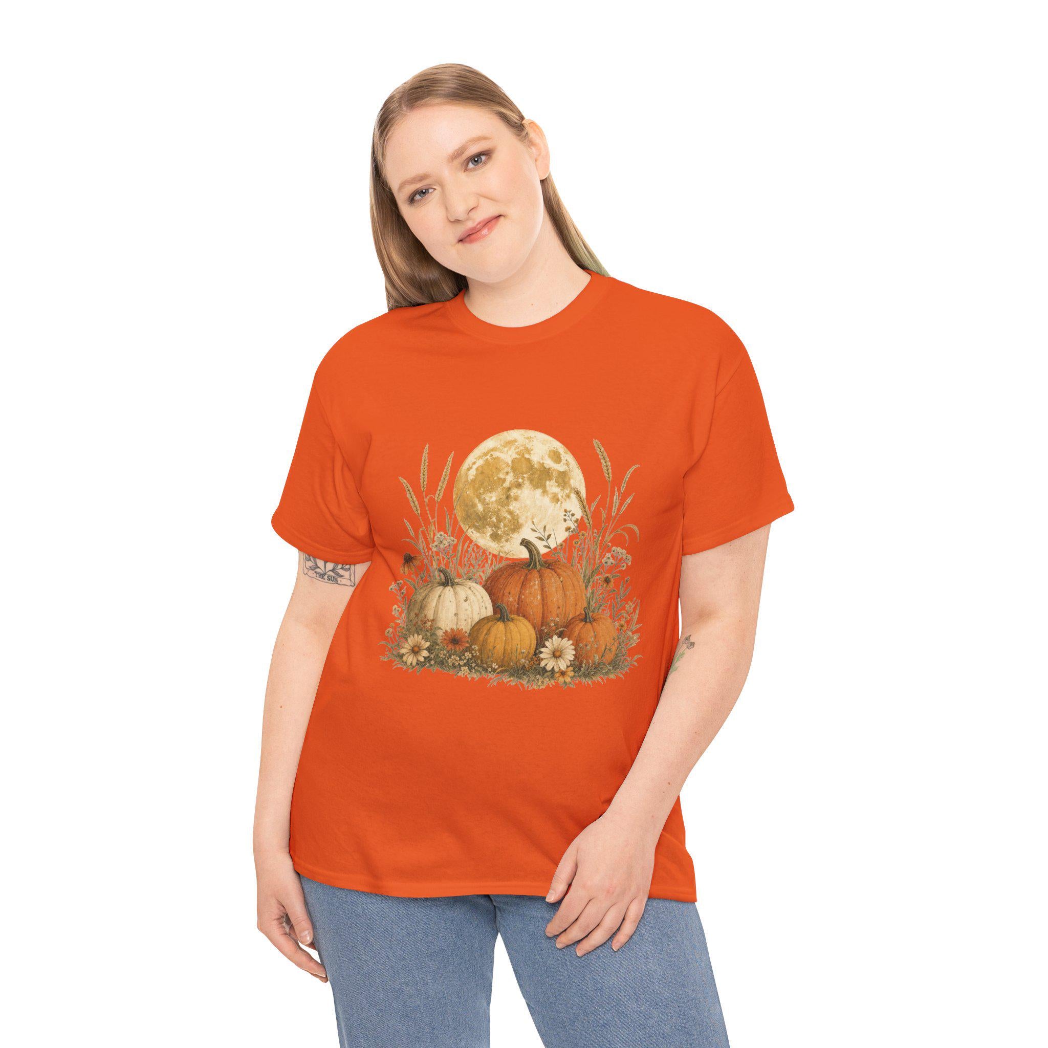 A woman models an orange t-shirt featuring a graphic of a large full moon behind a cluster of pumpkins, tall grasses, and small flowers.