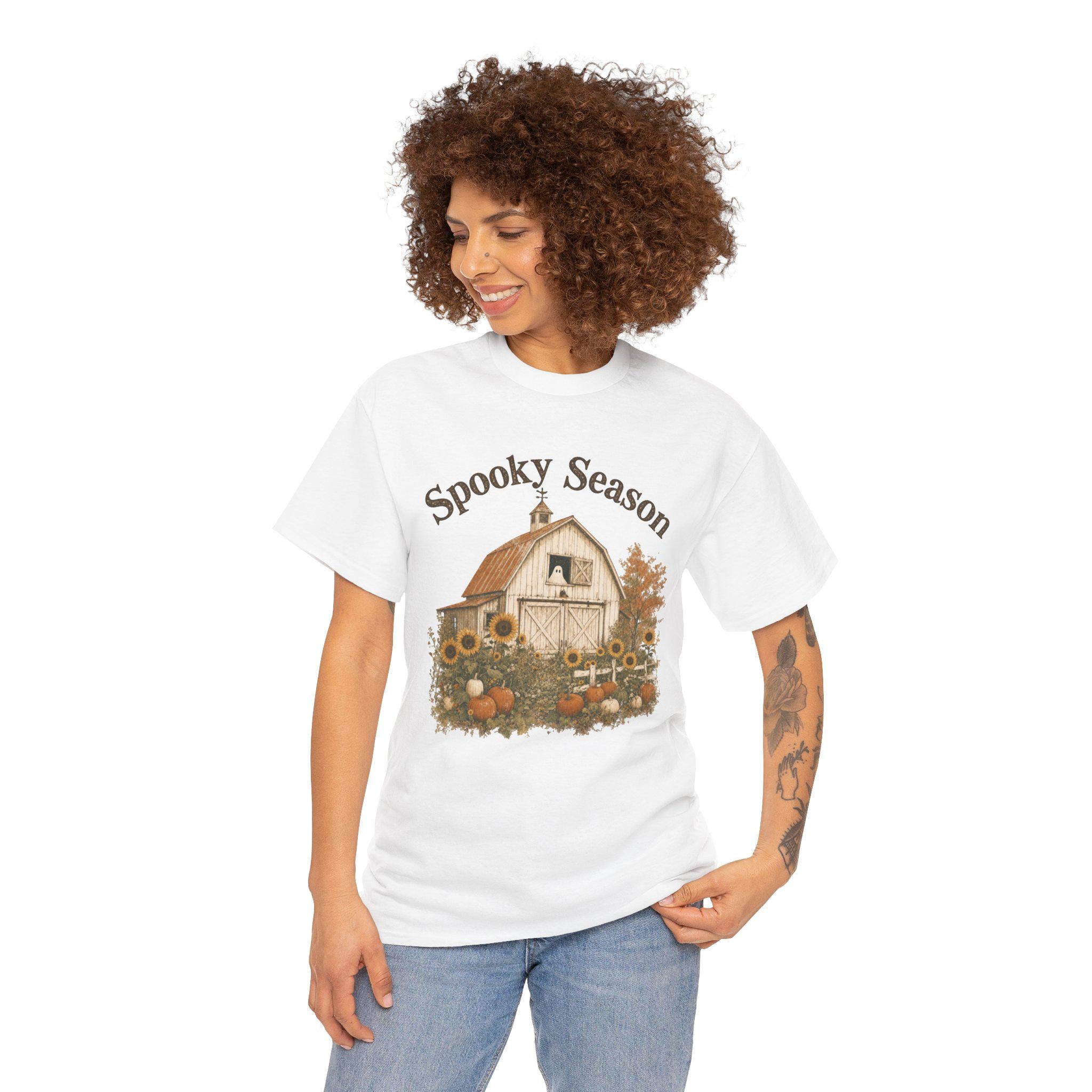 A smiling woman with curly hair wears a white t-shirt featuring a barn, sunflowers, pumpkins, a ghost in a window, and "Spooky Season" text.