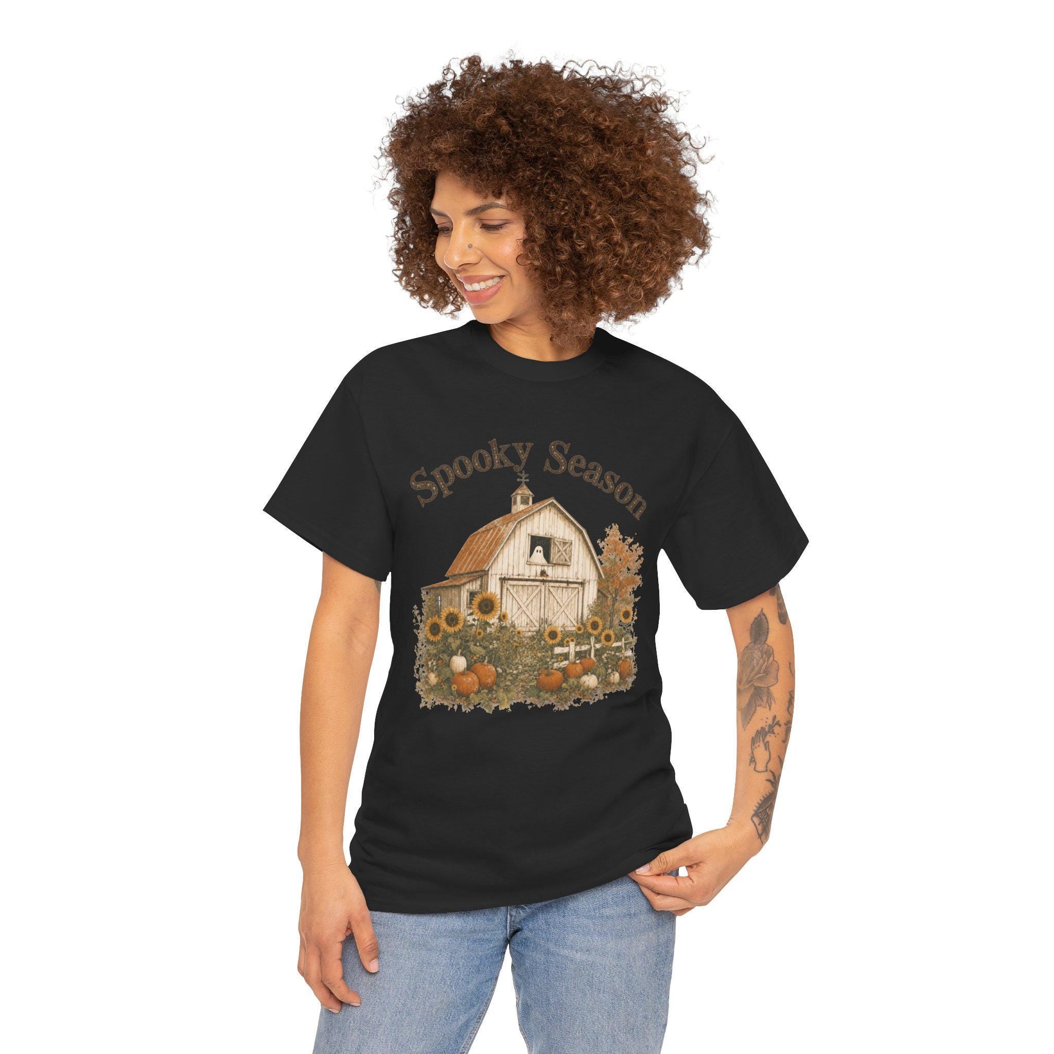 A smiling woman with curly hair wears a black t-shirt featuring a barn, sunflowers, pumpkins, a ghost in a window, and "Spooky Season" text.