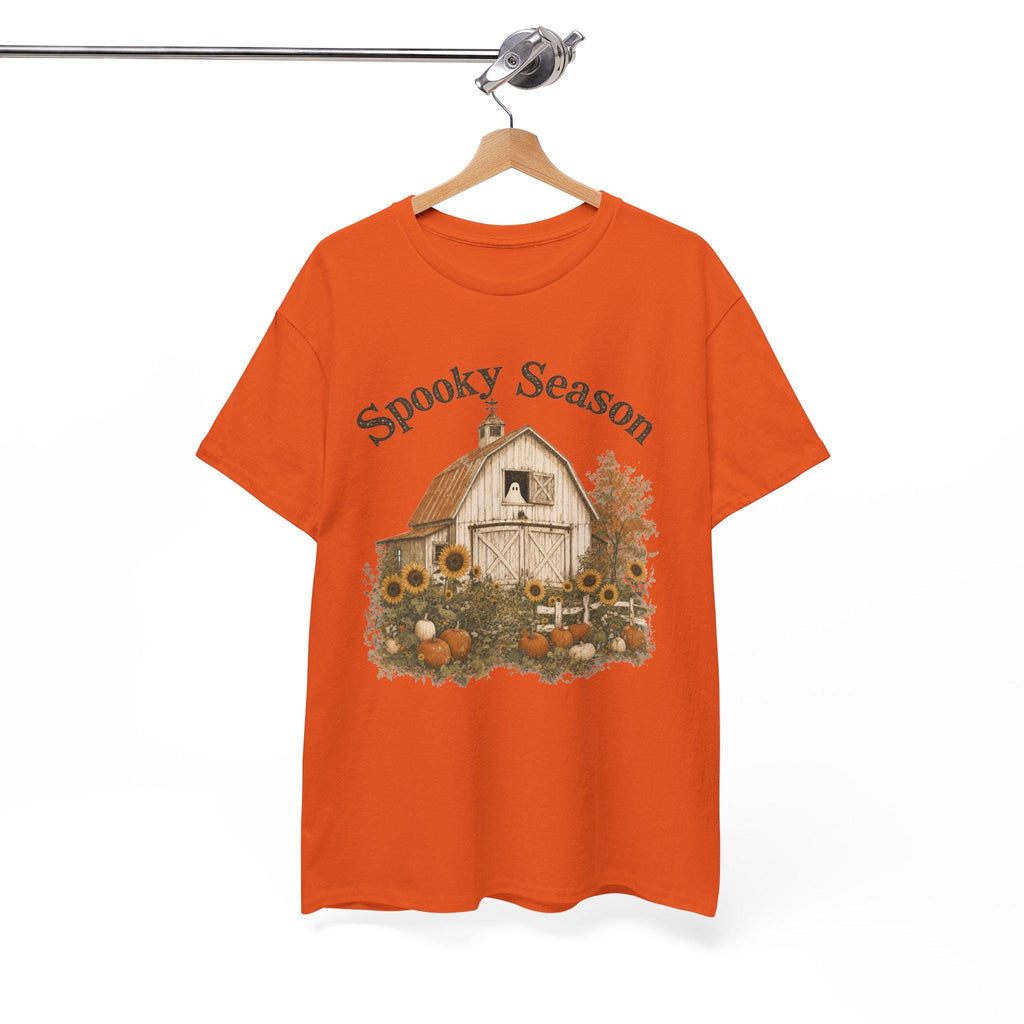 An orange t-shirt hanging on a wooden hanger, displaying a graphic of a white barn, sunflowers, and pumpkins, with "Spooky Season" text above.