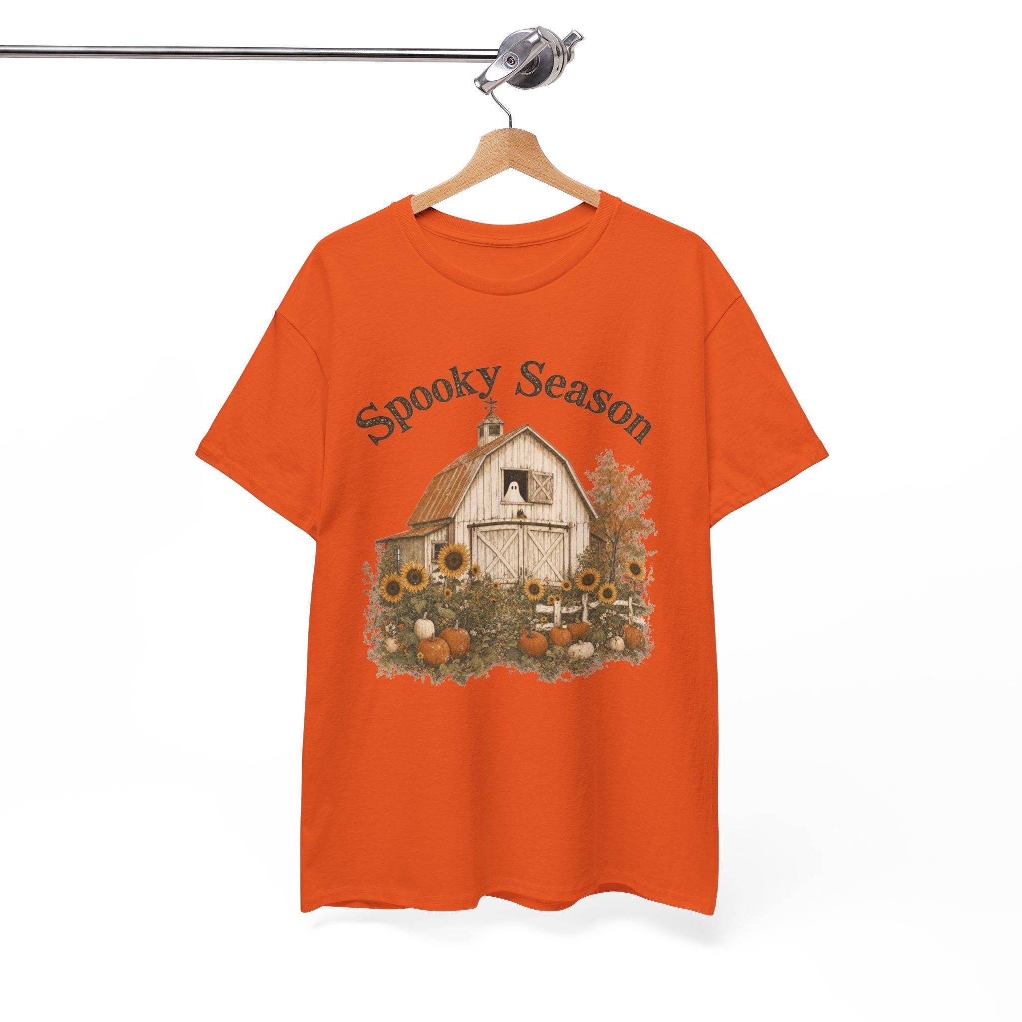 An orange t-shirt hanging on a wooden hanger, displaying a graphic of a white barn, sunflowers, and pumpkins, with "Spooky Season" text above.