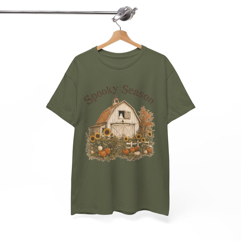 An olive green t-shirt hanging on a wooden hanger, displaying a graphic of a white barn, sunflowers, and pumpkins, with "Spooky Season" text above.
