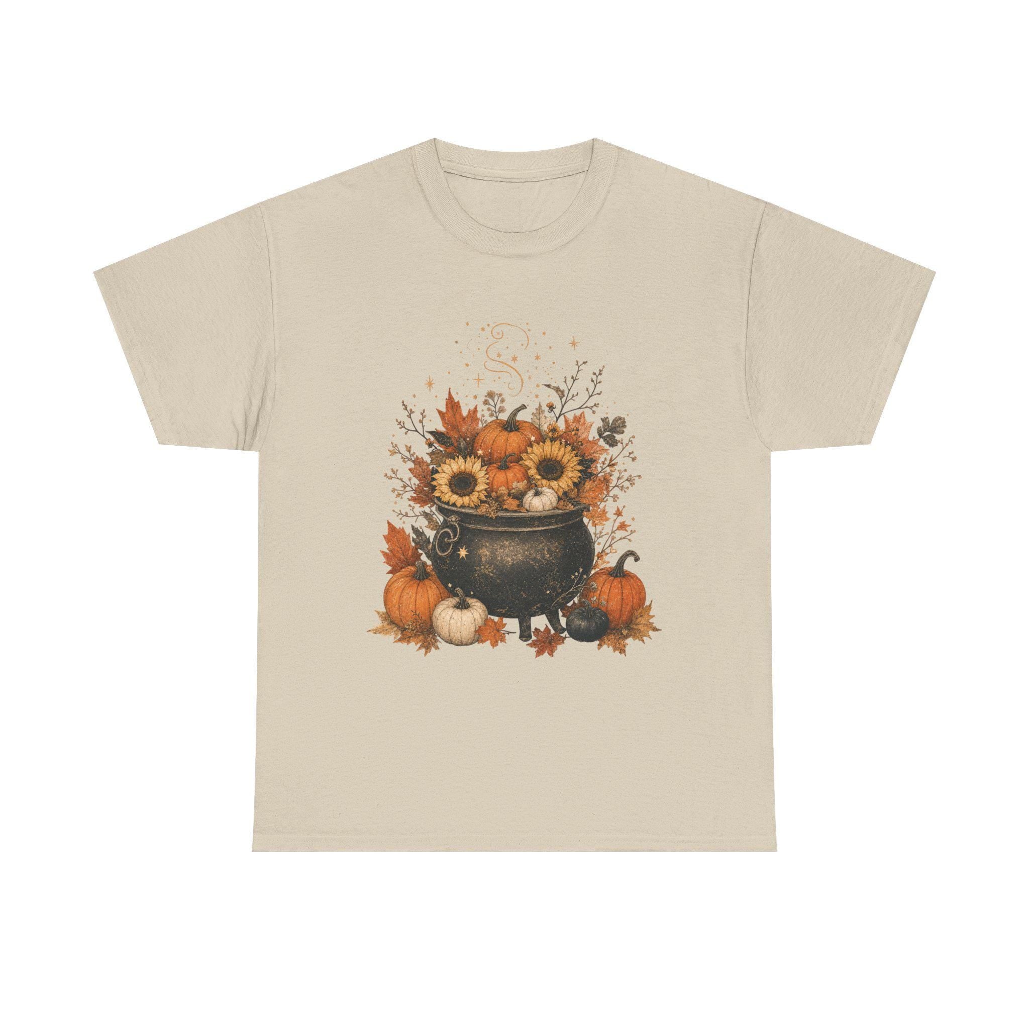 A light tan short-sleeved t-shirt with a fall-themed graphic of a black cauldron filled with sunflowers, pumpkins, and autumn leaves.