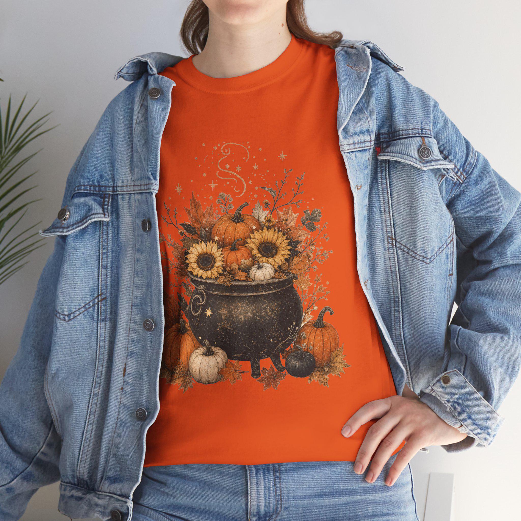 A woman in a denim jacket wears an orange t-shirt with a graphic of a black cauldron filled with pumpkins, sunflowers, and autumn leaves.