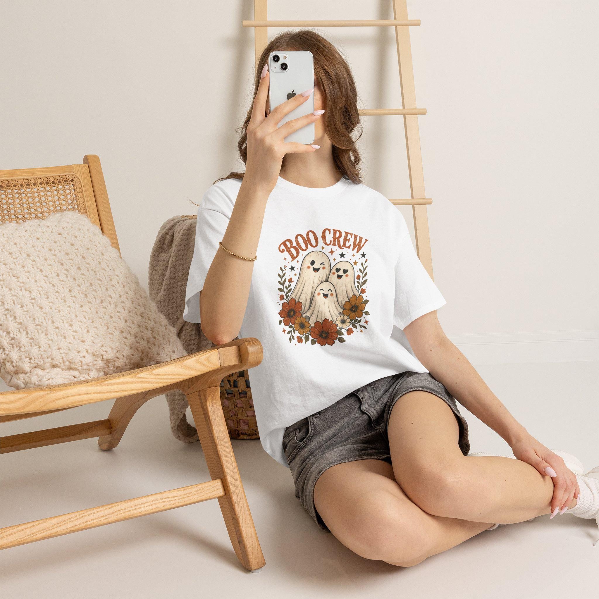 A person wearing a white t-shirt with a "Boo Crew" graphic featuring three ghosts surrounded by orange and white flowers, sitting on a light-colored floor.