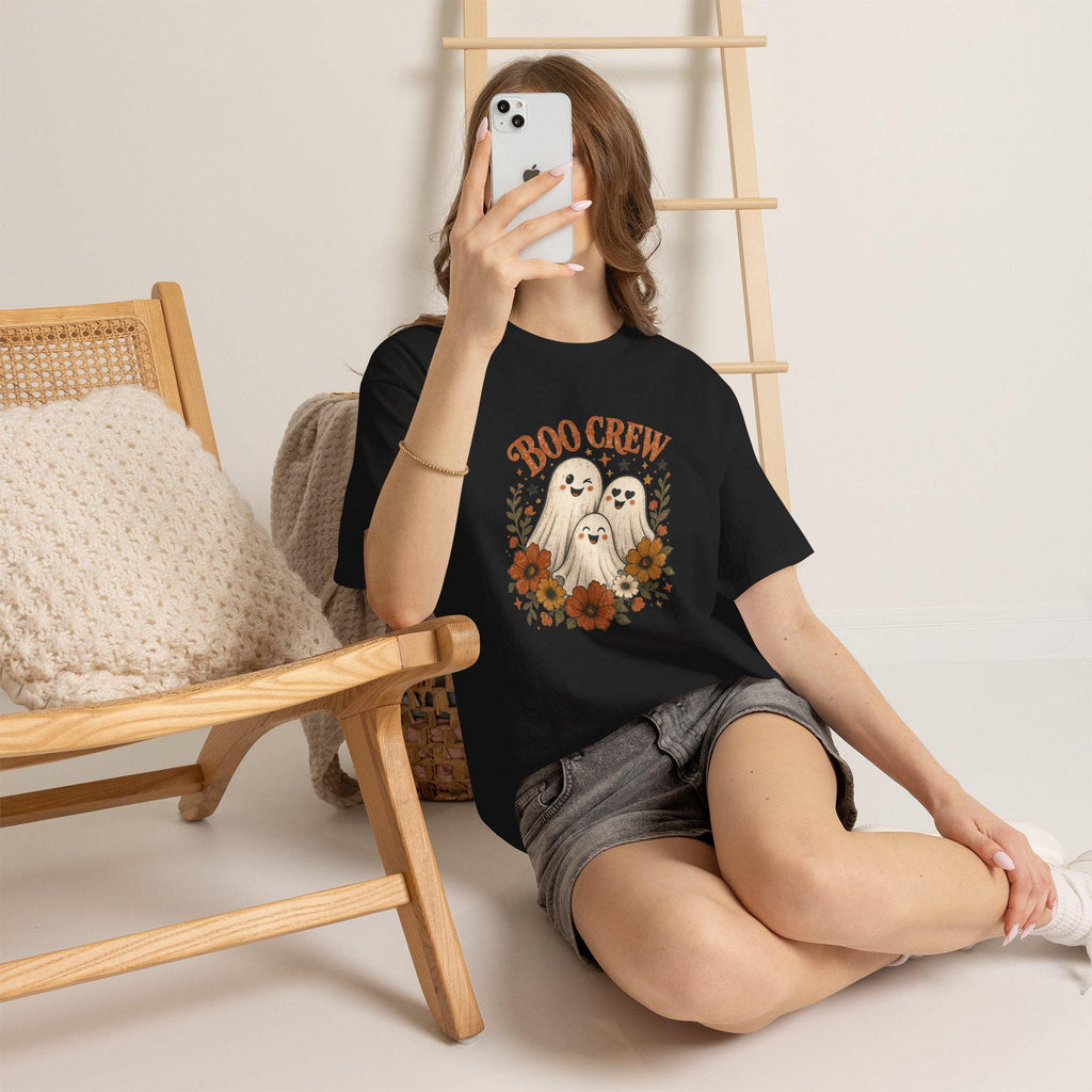 A person wearing a black t-shirt with a "Boo Crew" graphic featuring three ghosts surrounded by orange and white flowers, sitting on a light-colored floor.