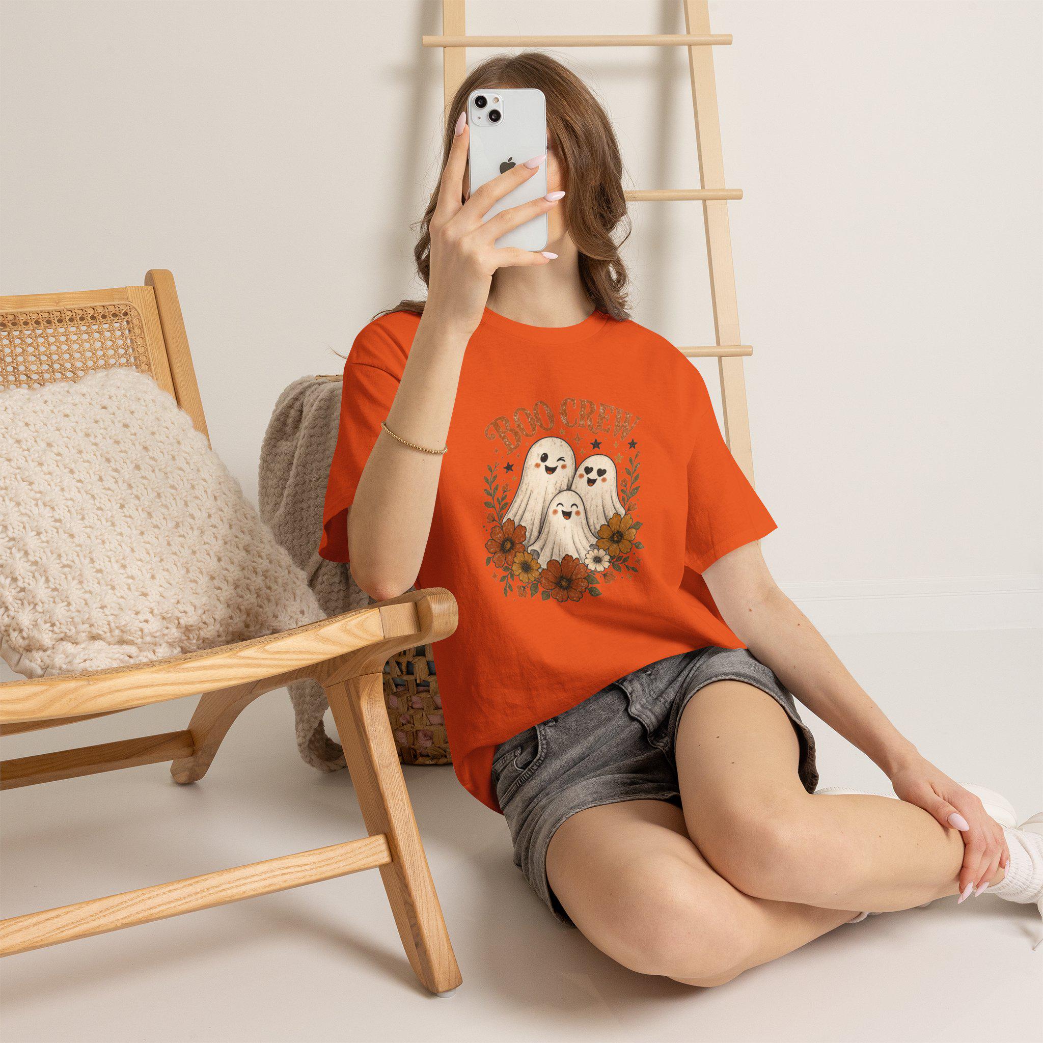 A person wearing an orange t-shirt with a "Boo Crew" graphic featuring three ghosts surrounded by orange and white flowers, sitting on a light-colored floor.