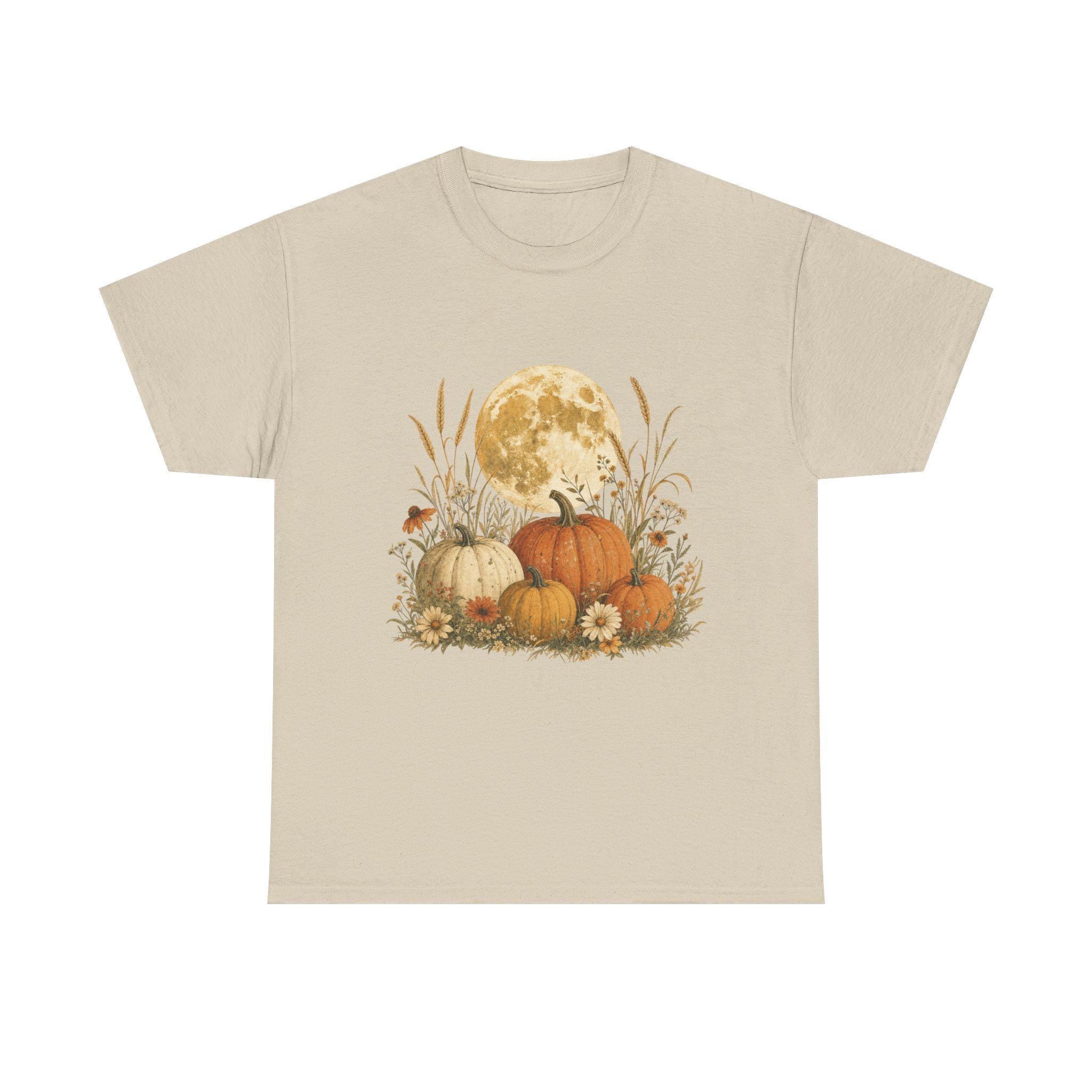 A light beige t-shirt with a graphic of a full moon behind a patch of pumpkins, tall grass, and flowers.
