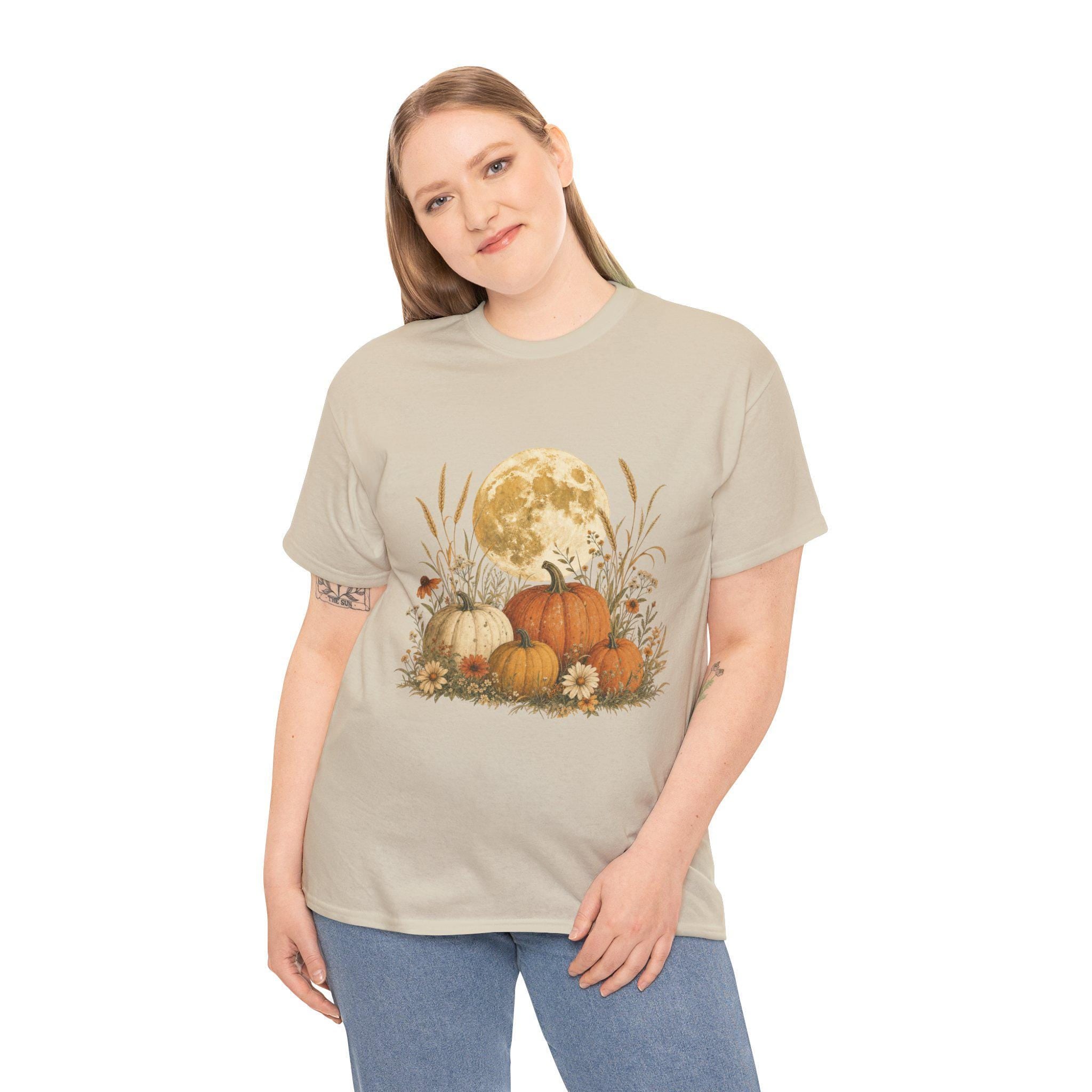 A woman with blonde hair wearing a light beige t-shirt with a graphic of a full moon behind a patch of pumpkins, tall grass, and flowers.