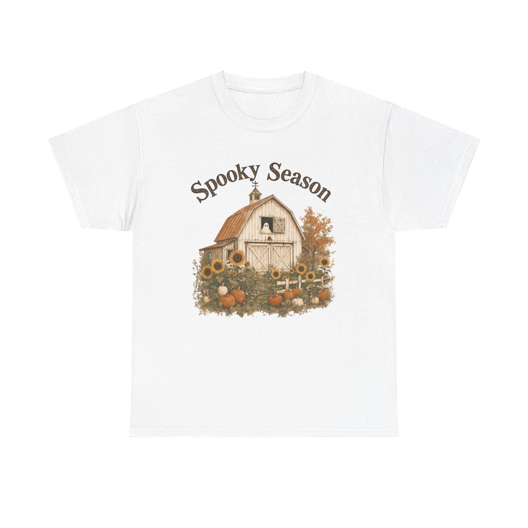 A white t-shirt with a graphic of a barn, sunflowers, pumpkins, and a ghost in a window, with "Spooky Season" text.