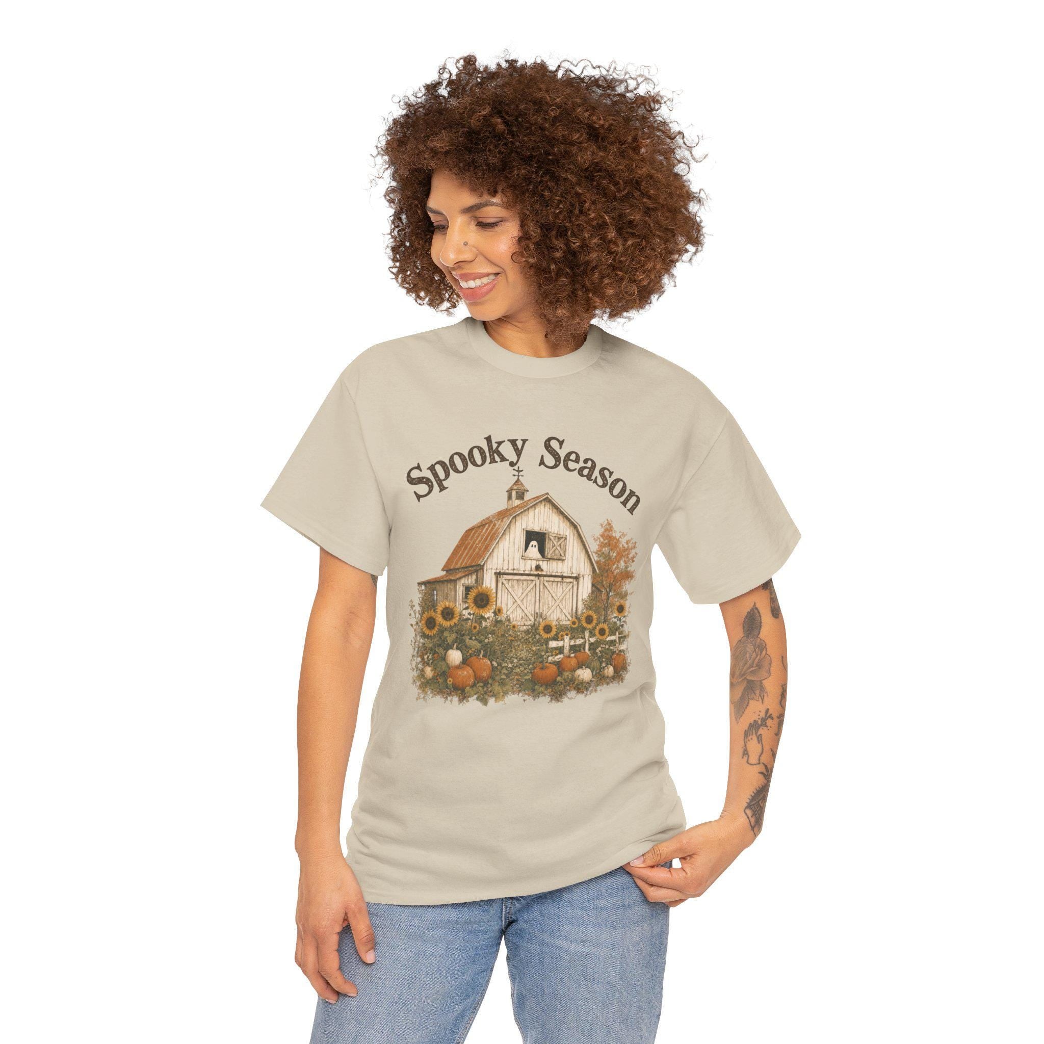A smiling woman with curly hair wears a light tan t-shirt featuring a barn, sunflowers, pumpkins, a ghost in a window, and "Spooky Season" text.