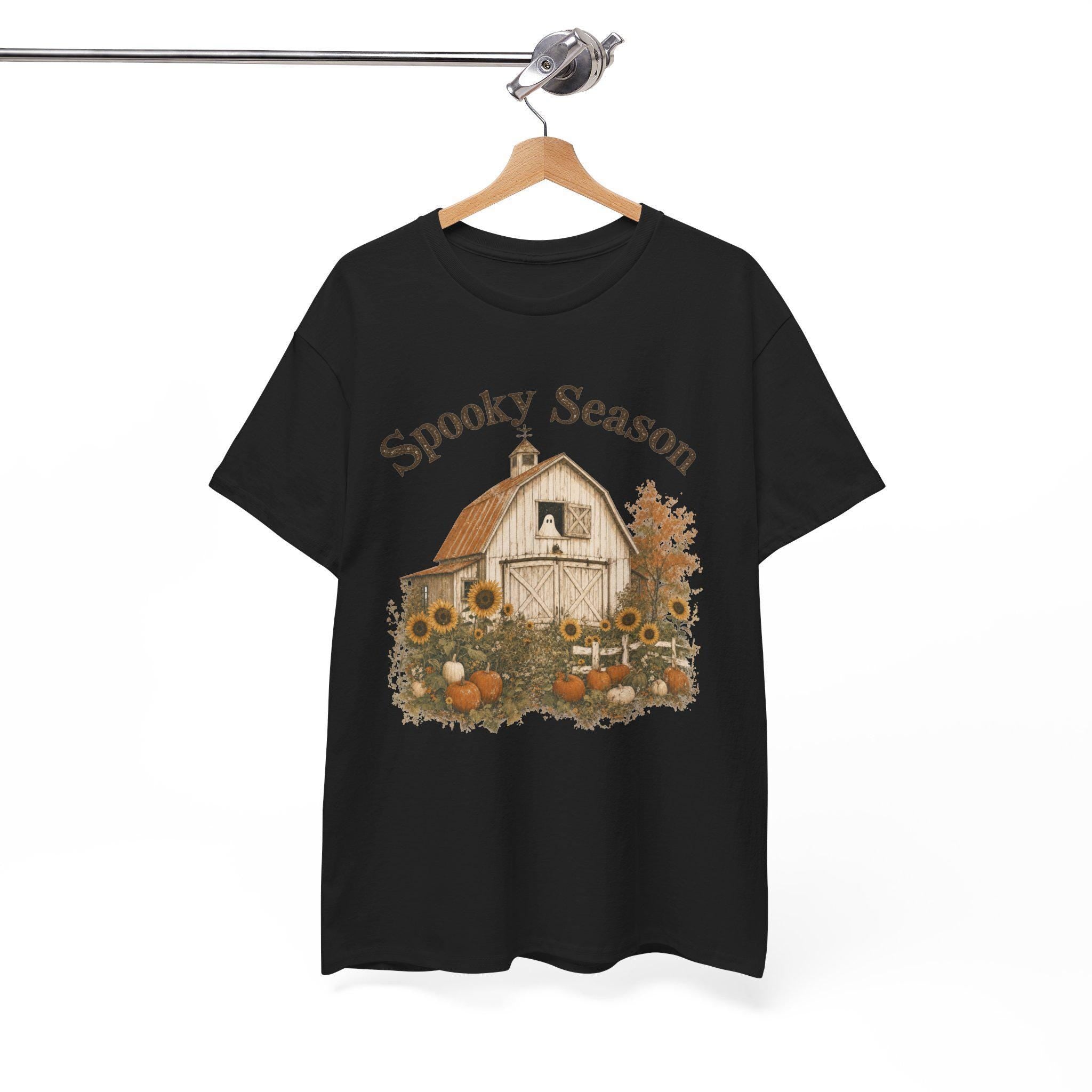 A black t-shirt hanging on a wooden hanger, displaying a graphic of a white barn, sunflowers, and pumpkins, with "Spooky Season" text above.