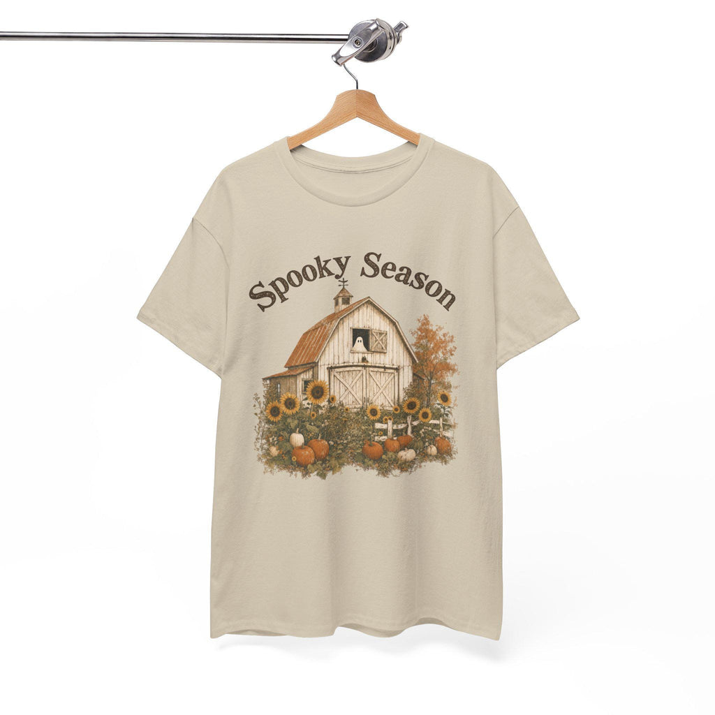 A light beige t-shirt hanging on a wooden hanger, displaying a graphic of a white barn, sunflowers, and pumpkins, with "Spooky Season" text above.