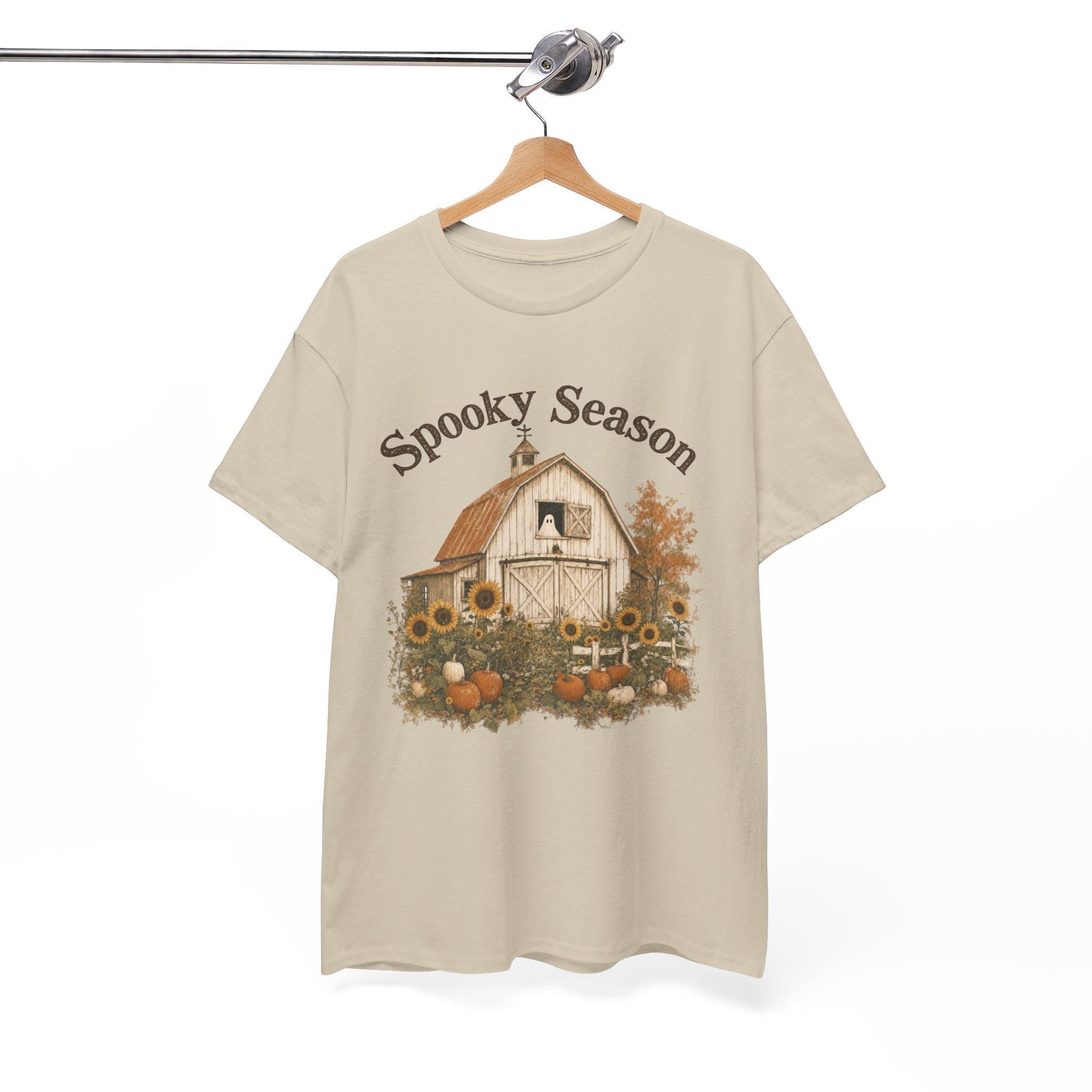 A light beige t-shirt hanging on a wooden hanger, displaying a graphic of a white barn, sunflowers, and pumpkins, with "Spooky Season" text above.