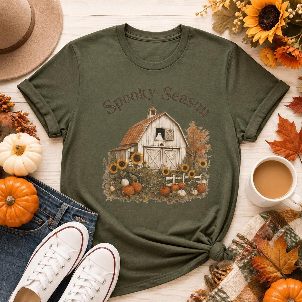 An olive green t-shirt with a barn, sunflowers, pumpkins, and a ghost in a window, with "Spooky Season" text, surrounded by fall decor.