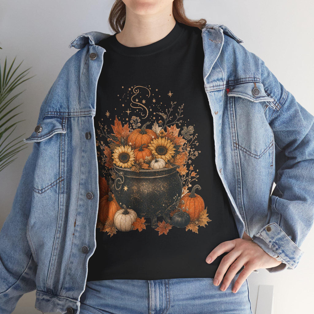 A woman in a denim jacket wears a black t-shirt with a graphic of a black cauldron filled with pumpkins, sunflowers, and autumn leaves.