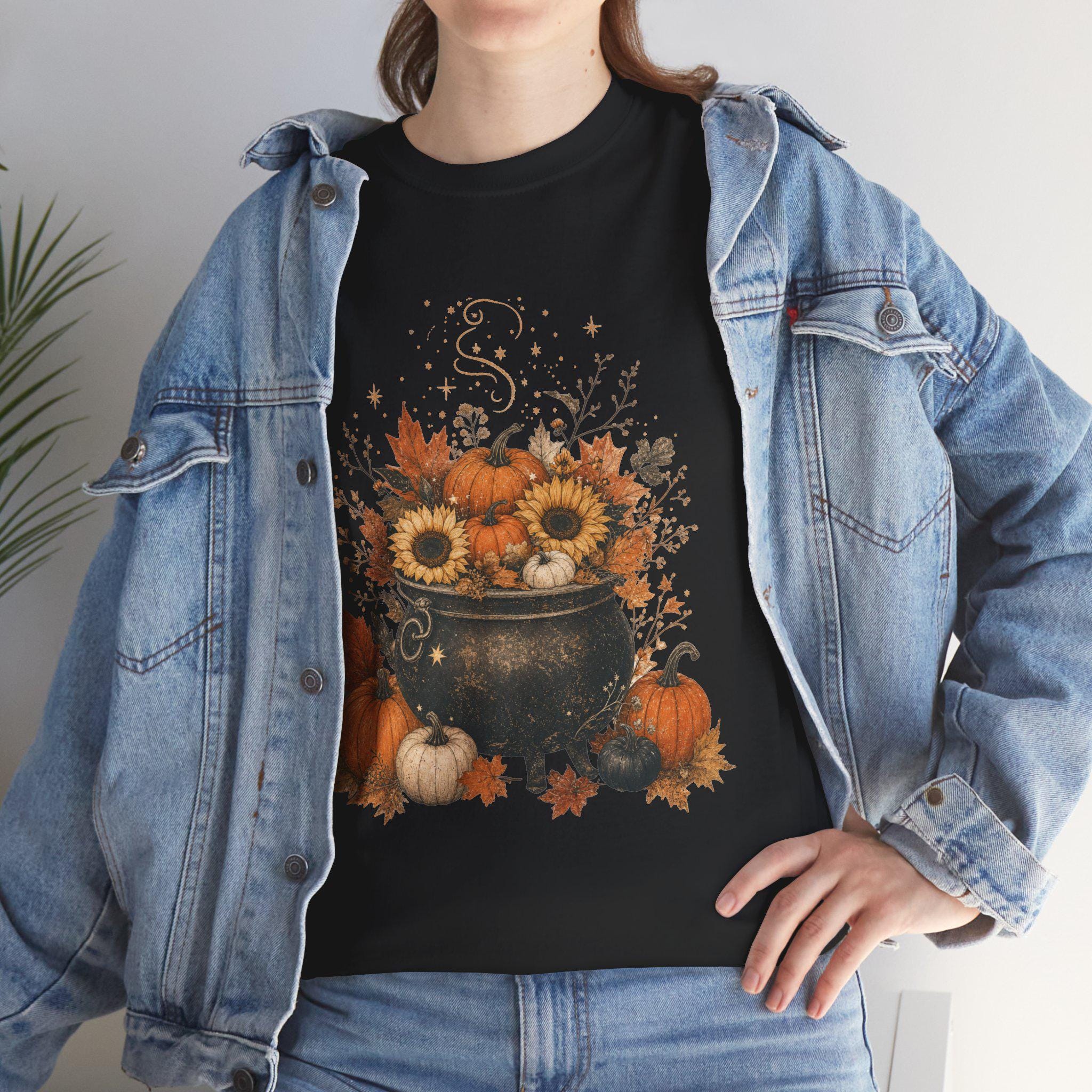 A woman in a denim jacket wears a black t-shirt with a graphic of a black cauldron filled with pumpkins, sunflowers, and autumn leaves.