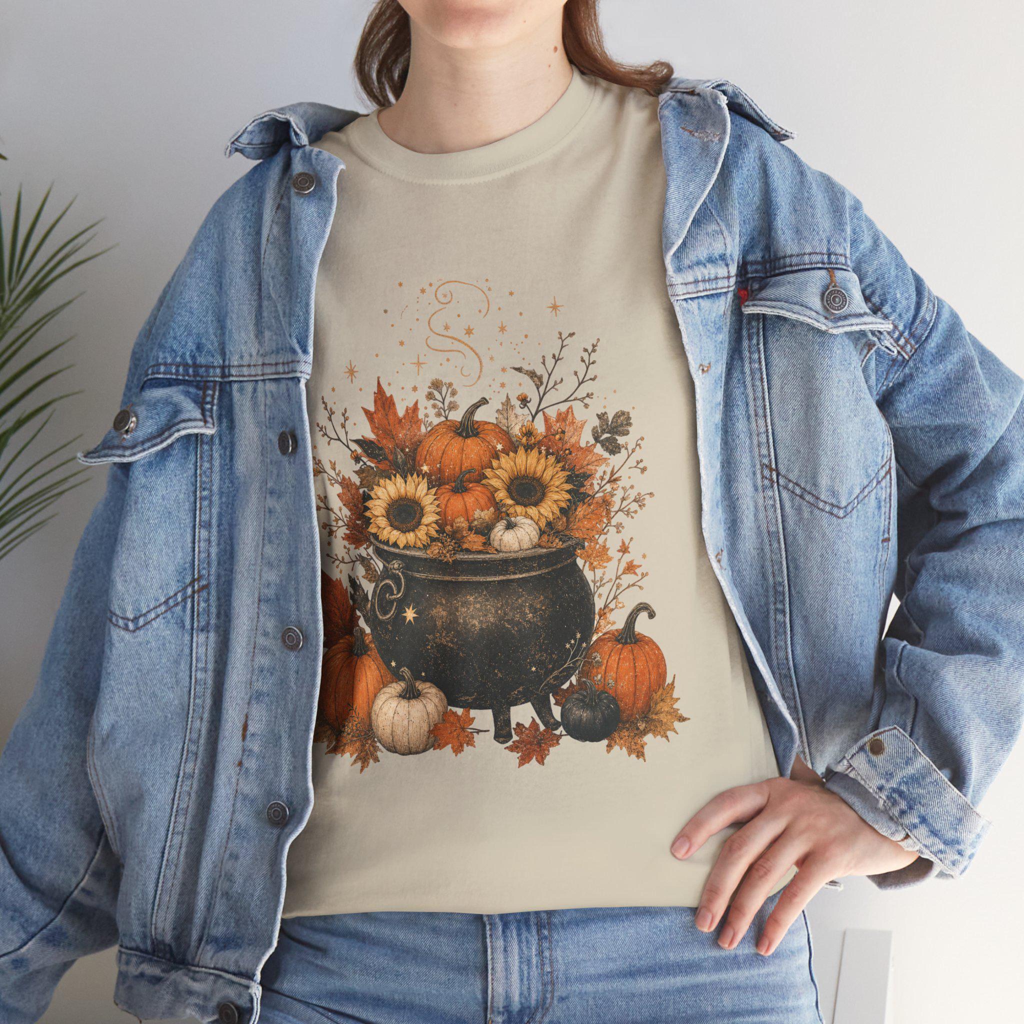 A woman in a denim jacket wears a light tan t-shirt with a graphic of a black cauldron filled with pumpkins, sunflowers, and autumn leaves.