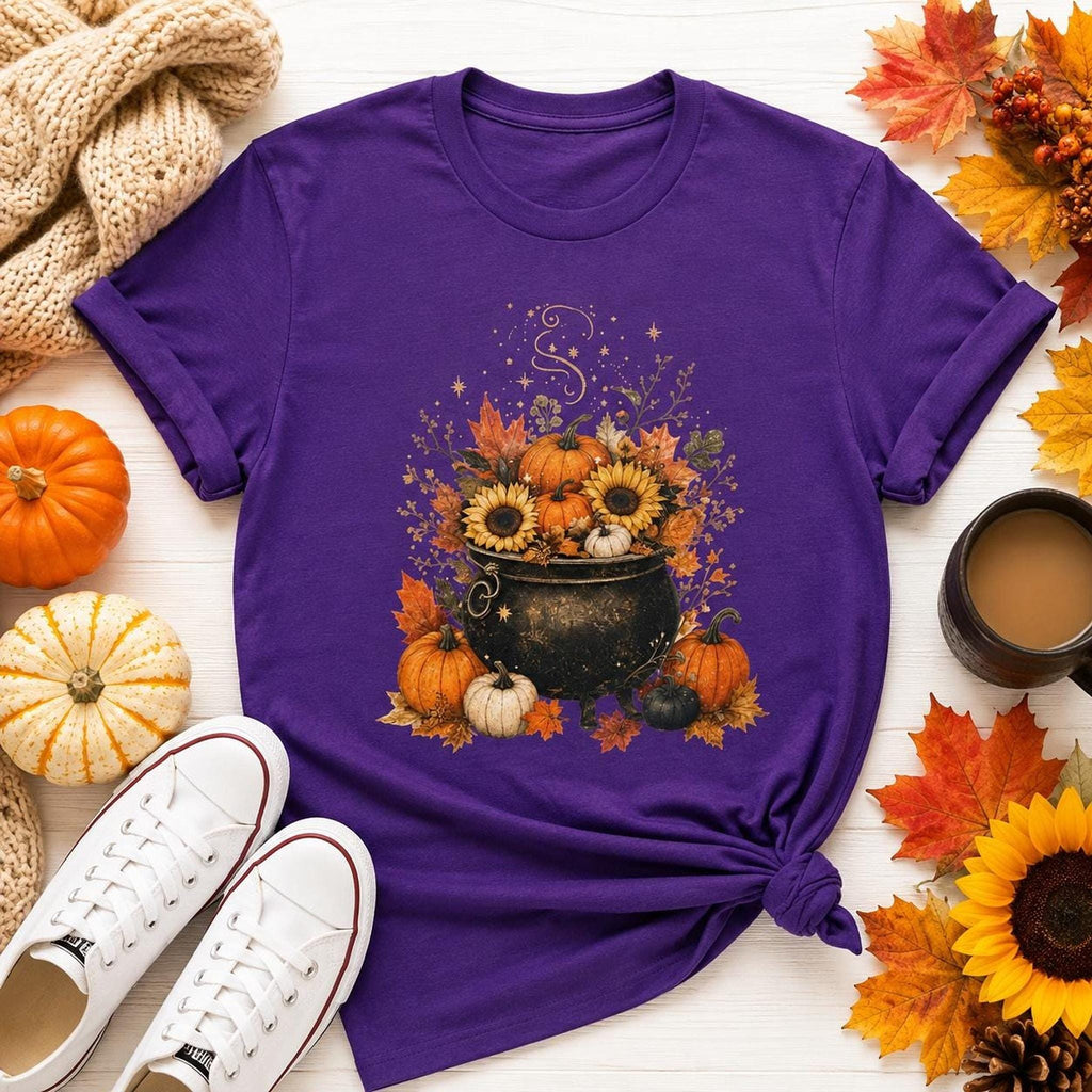 A purple short-sleeved t-shirt with a fall-themed graphic of a black cauldron filled with sunflowers, pumpkins, and autumn leaves, surrounded by real pumpkins, leaves, and white sneakers.