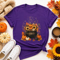 A purple short-sleeved t-shirt with a fall-themed graphic of a black cauldron filled with sunflowers, pumpkins, and autumn leaves, surrounded by real pumpkins, leaves, and white sneakers.