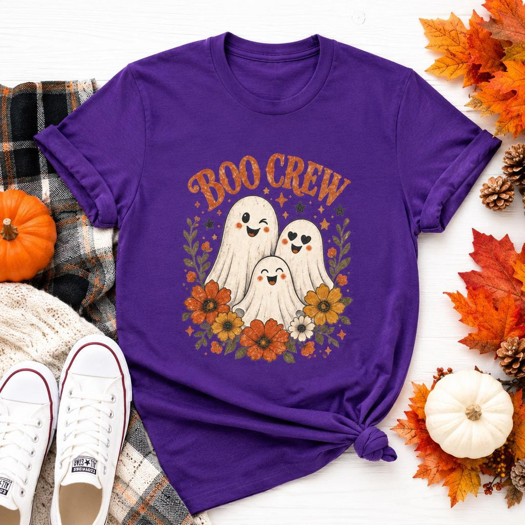 A purple t-shirt with a "Boo Crew" graphic featuring three ghosts surrounded by orange and white flowers, displayed on a wooden surface with fall leaves and a small pumpkin.
