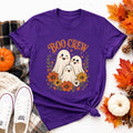 A purple t-shirt with a "Boo Crew" graphic featuring three ghosts surrounded by orange and white flowers, displayed on a wooden surface with fall leaves and a small pumpkin.