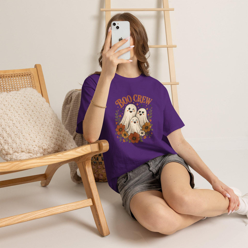 A person with light skin and brown hair sits on a light wooden floor, holding a phone to their face, wearing a purple t-shirt with "BOO CREW" and three smiling ghosts surrounded by orange and brown flowers.